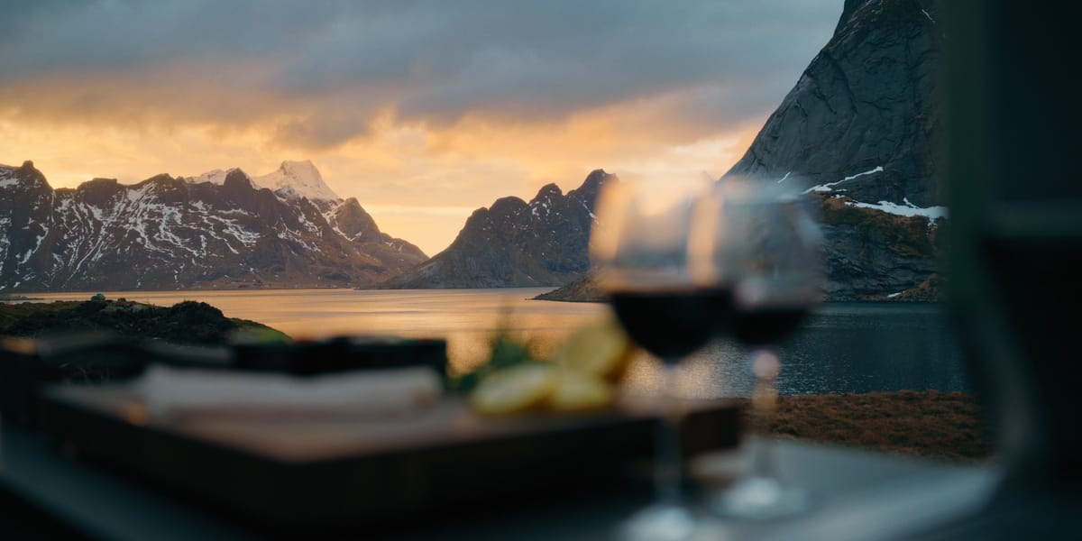 Two glasses of red wine and a cutting board on a dark stone countertop, a fjord and mountains at sunset behind them