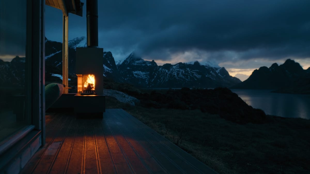 A FREILUFT Fire burning under the roof of a wooden deck, a fjord and snowy mountains behind it at dusk