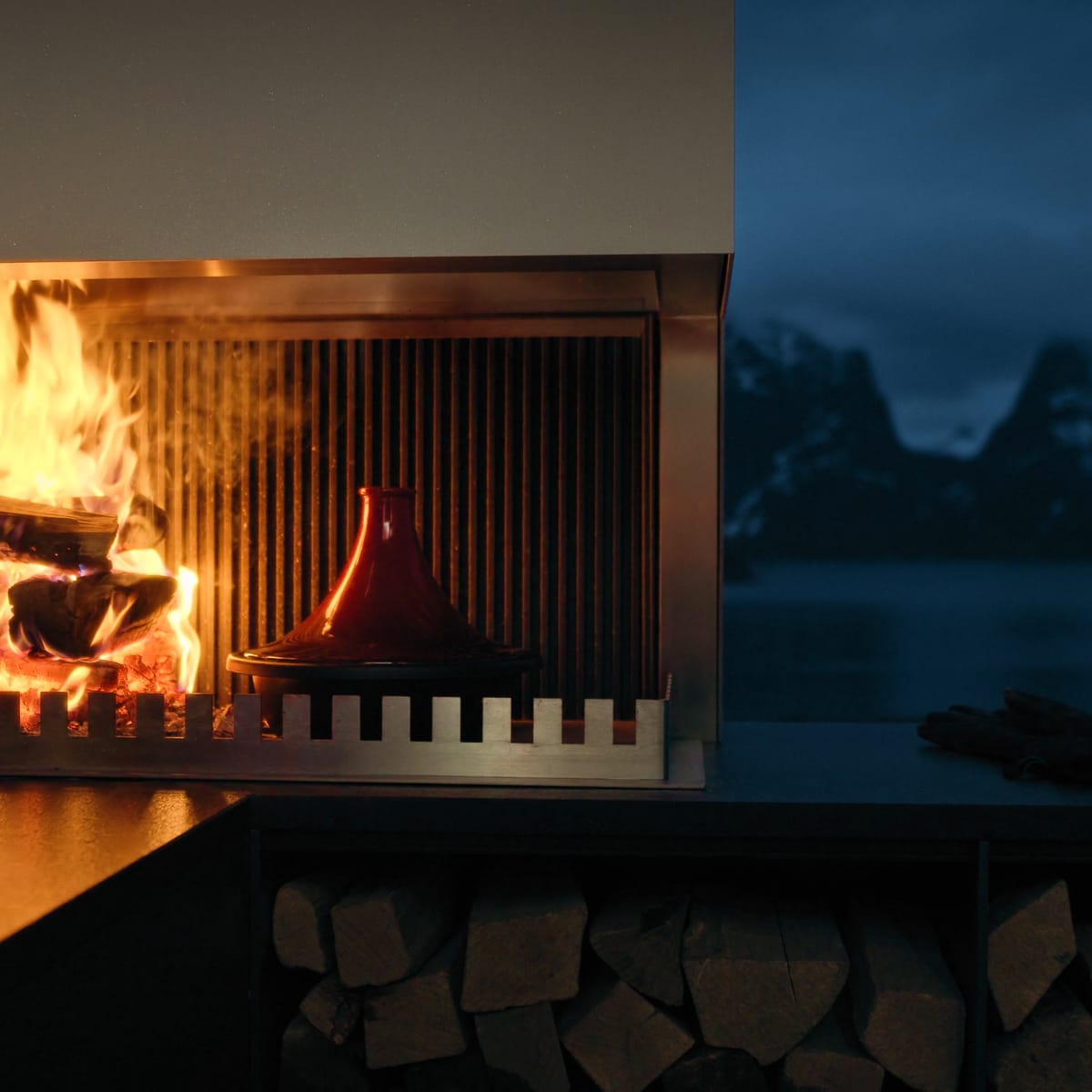 A FREILUFT grill-and-fire element with a burning wood fire and open log storage below, a fjord and snowy mountains behind it at dusk
