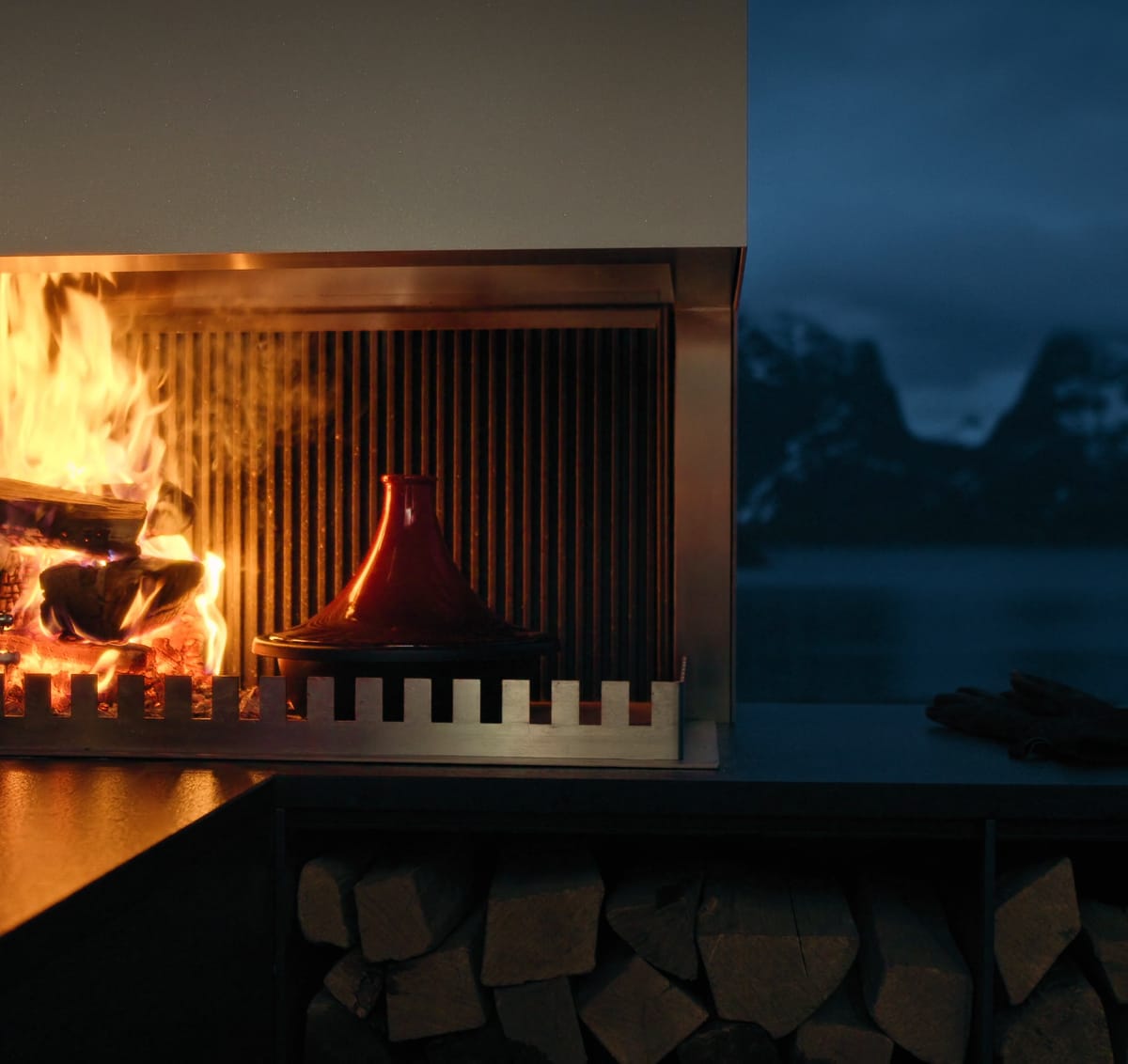 A FREILUFT grill-and-fire element with a burning wood fire and open log storage below, a fjord and snowy mountains behind it at dusk