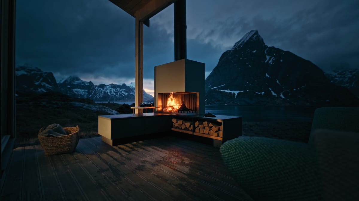 A FREILUFT Fire with a burning wood fire and log storage on a covered wooden deck, a fjord and mountains behind it at dusk