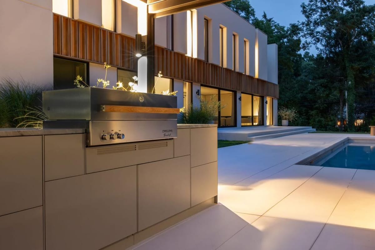 FREILUFT kitchen with a lit built-in grill beside the pool of a modern house on Long Island, New York, at dusk